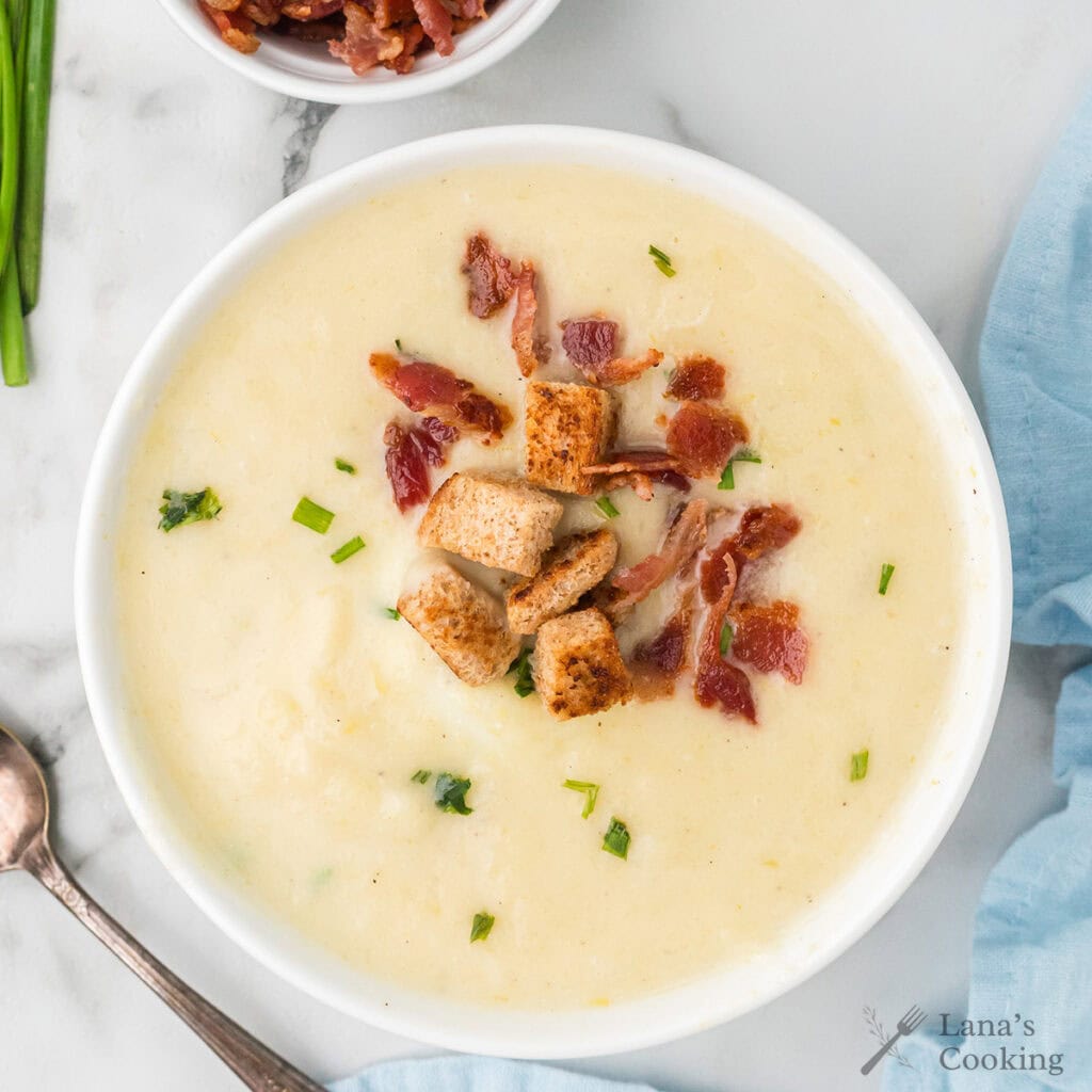 Bowl of creamy potato soup topped with croutons, bacon pieces, and chopped chives on a white marble surface.