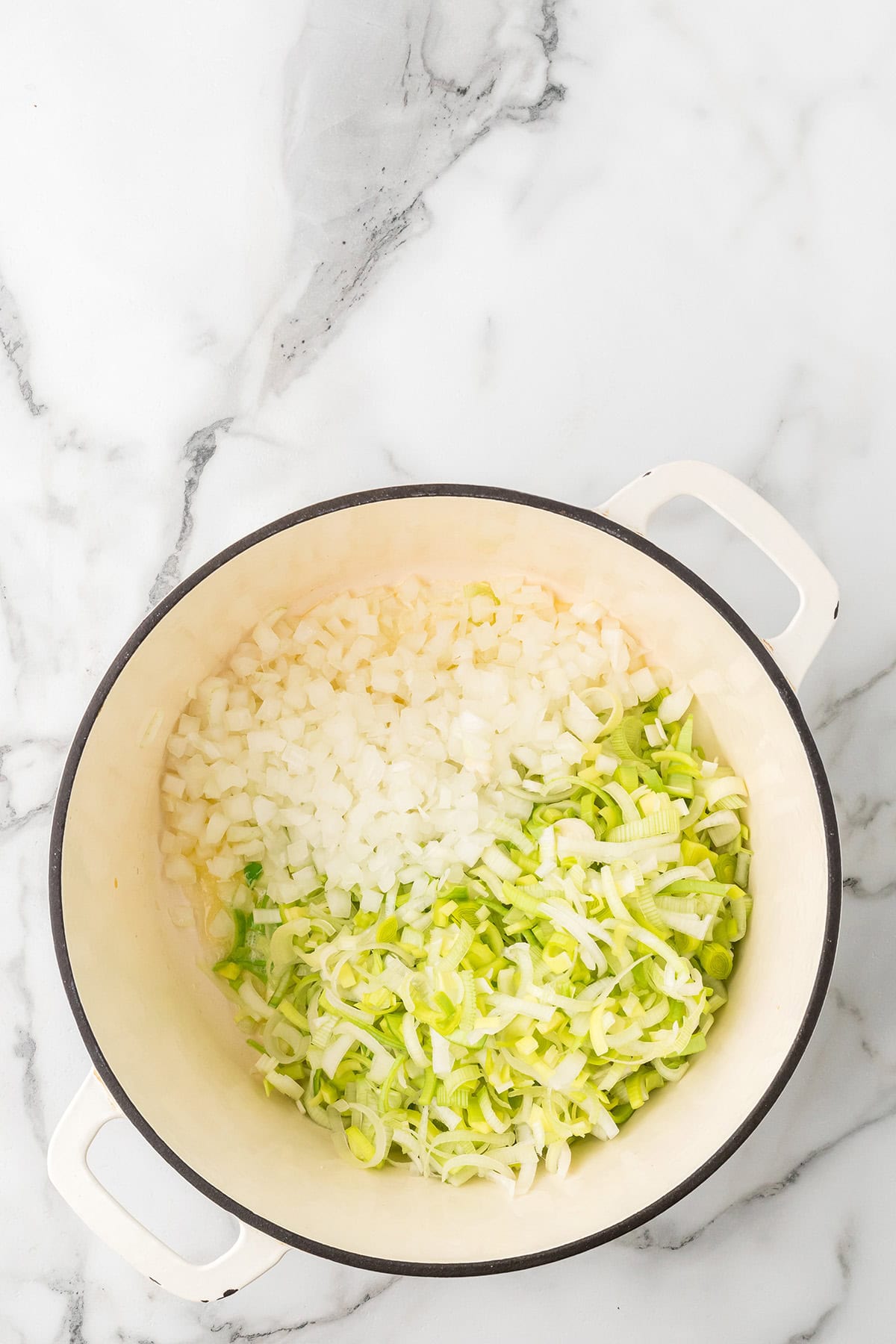 Chopped onions and leeks in a white pot on a marble countertop, ready for cooking.