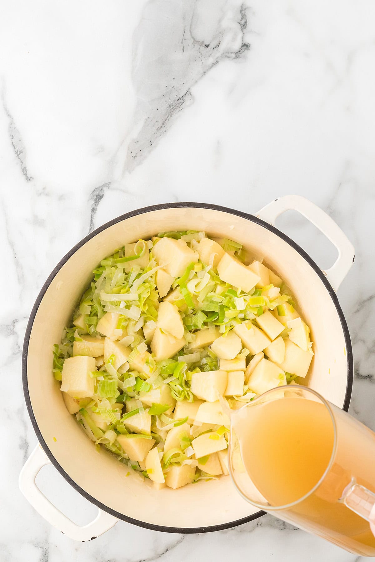 Diced potatoes and leeks in a white pot with broth being poured in on a marble surface.
