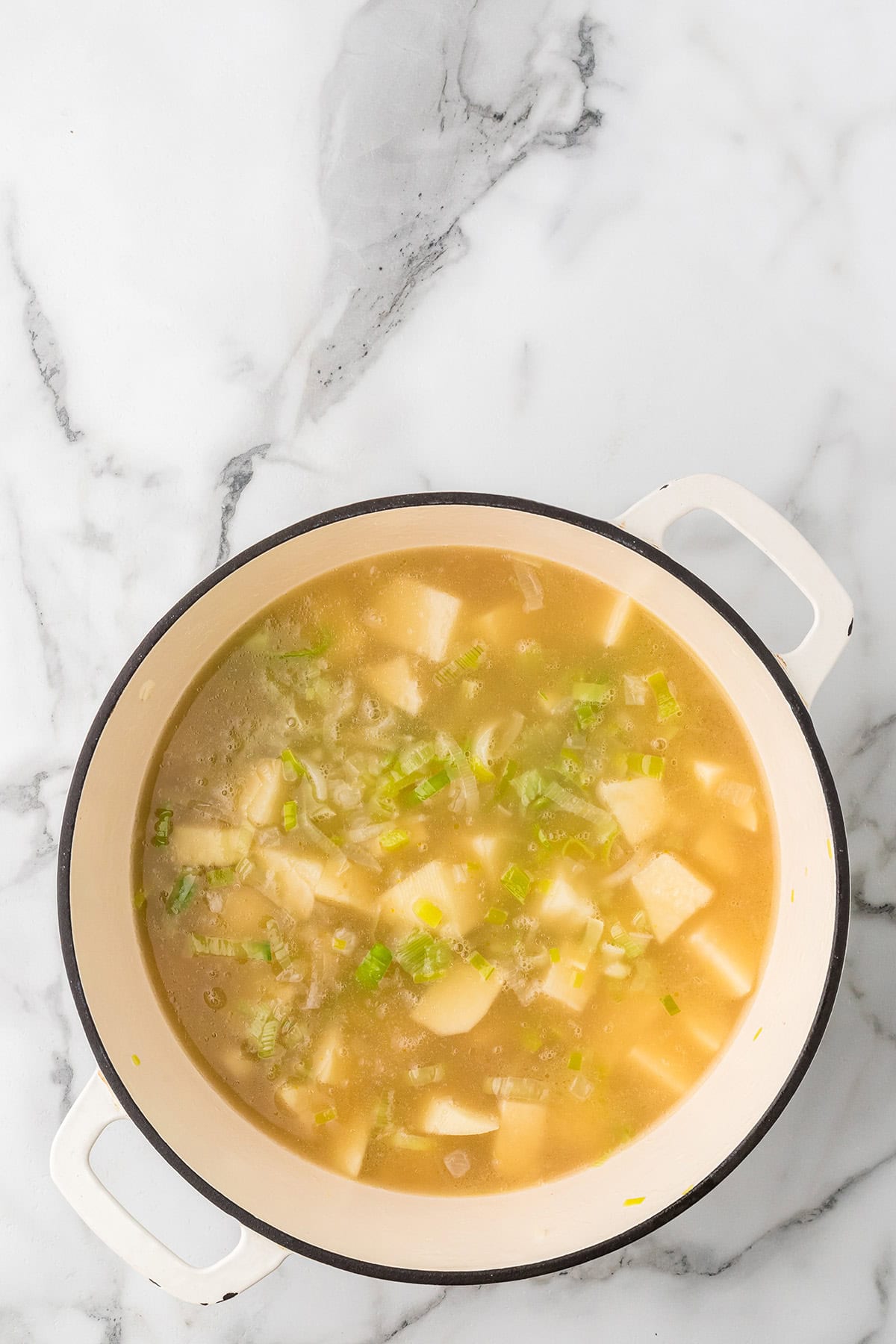 A pot of soup with diced potatoes and leeks on a white marble surface.