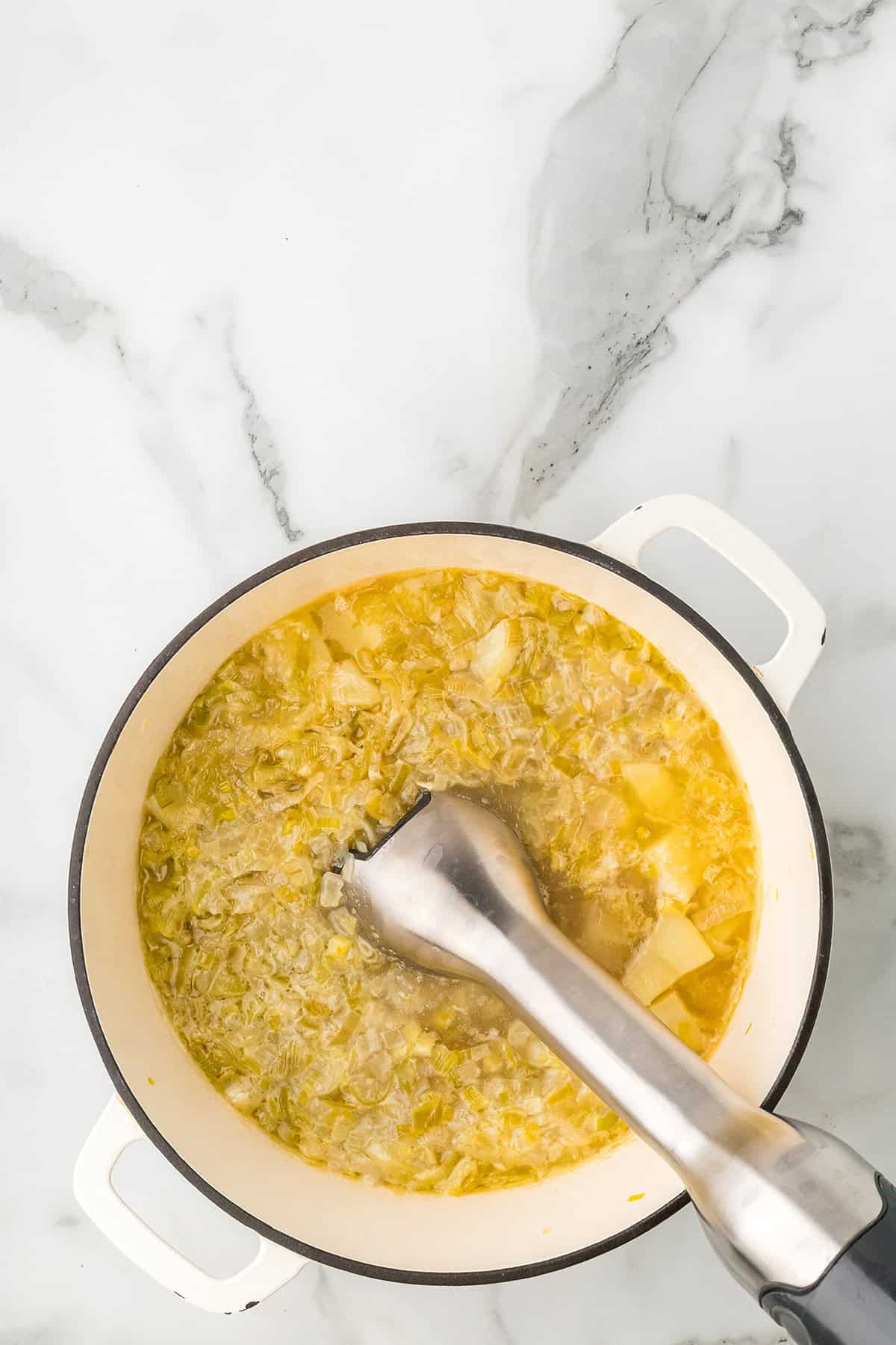 Overhead view of soup being blended in a white pot with an immersion blender on a marble surface.