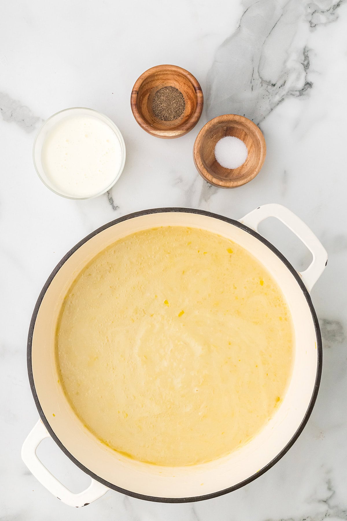 Overhead view of a pot of creamy soup with bowls of pepper, salt, and cream on a marble surface.