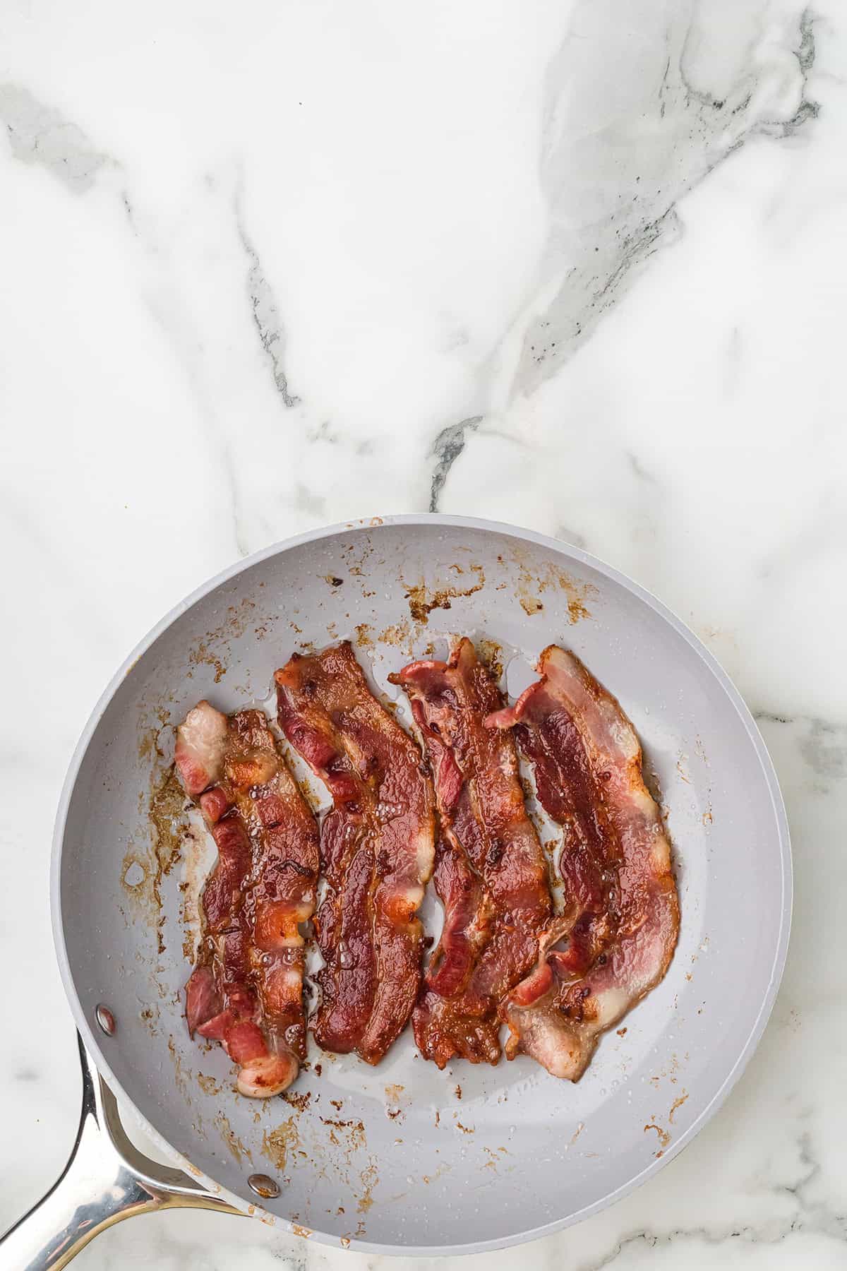 Four cooked bacon strips in a gray frying pan on a white marble countertop.