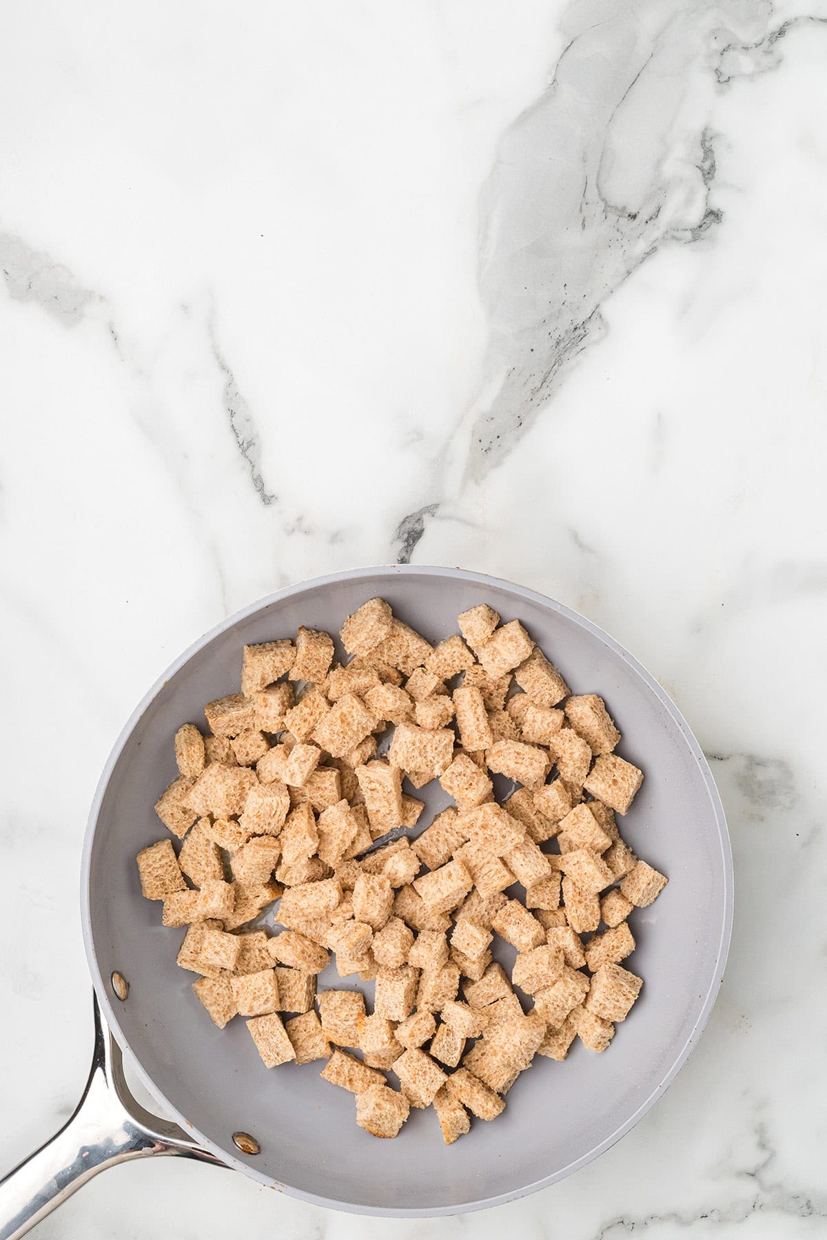 A frying pan filled with cubed bread pieces on a white marble surface.