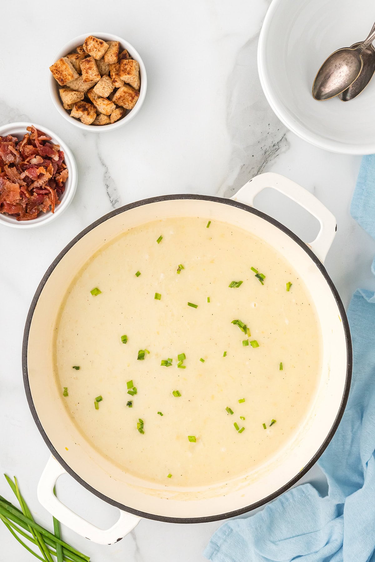 A pot of creamy soup garnished with chives, with bowls of bacon bits and croutons on the side.
