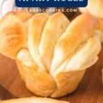 A golden-brown, homemade pull-apart roll sits on a wooden surface, with more rolls in the background.