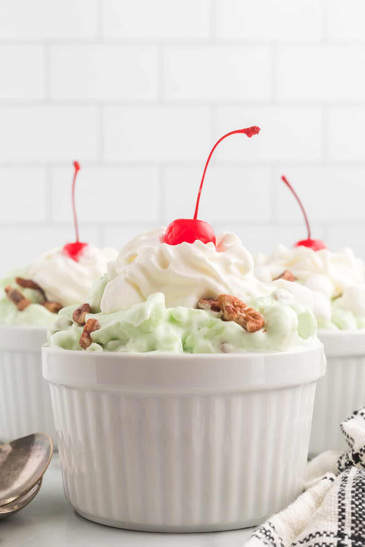 Small white bowls of green pistachio salad topped with whipped cream, a cherry, and walnuts.