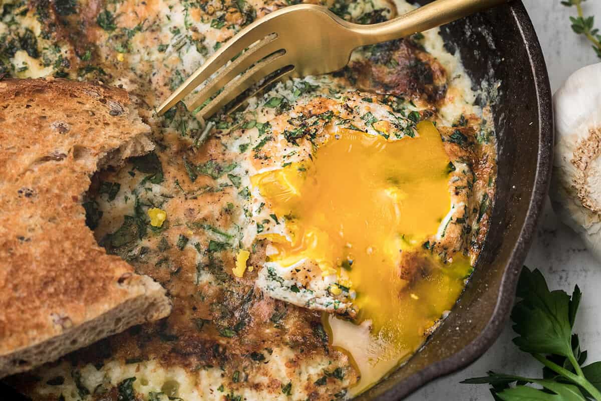 A fork and bread slice dip into a skillet of baked eggs with runny yolk, herbs, and cream.