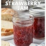Two jars of homemade strawberry jam with sliced bread in the background on a white surface.