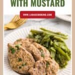 Plate of pork tenderloin with mustard next to green beans on a white plate, gold fork in the background.