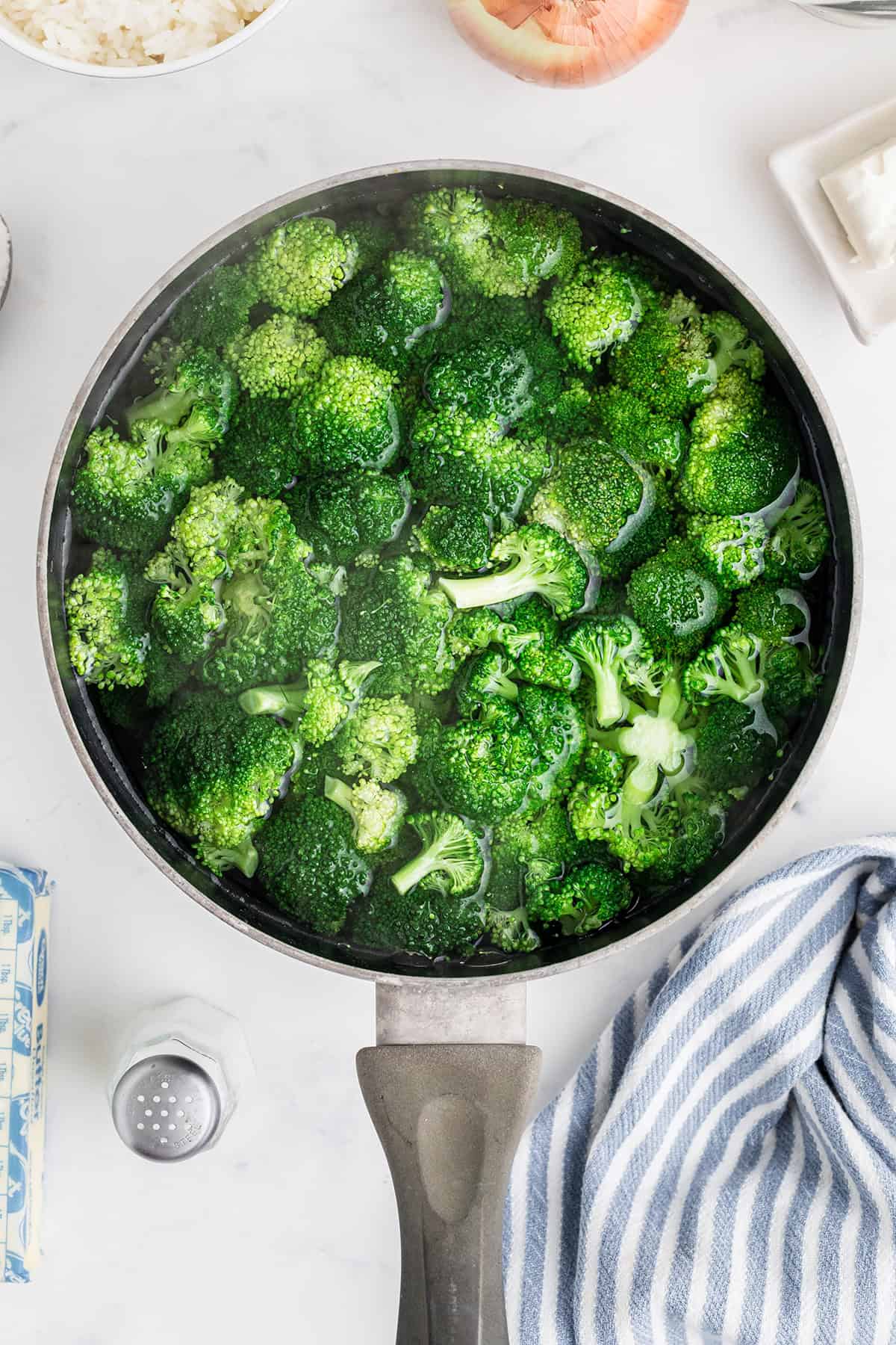 Broccoli florets boiling in water in a saucepan, with kitchen towel and ingredients nearby on a white surface.