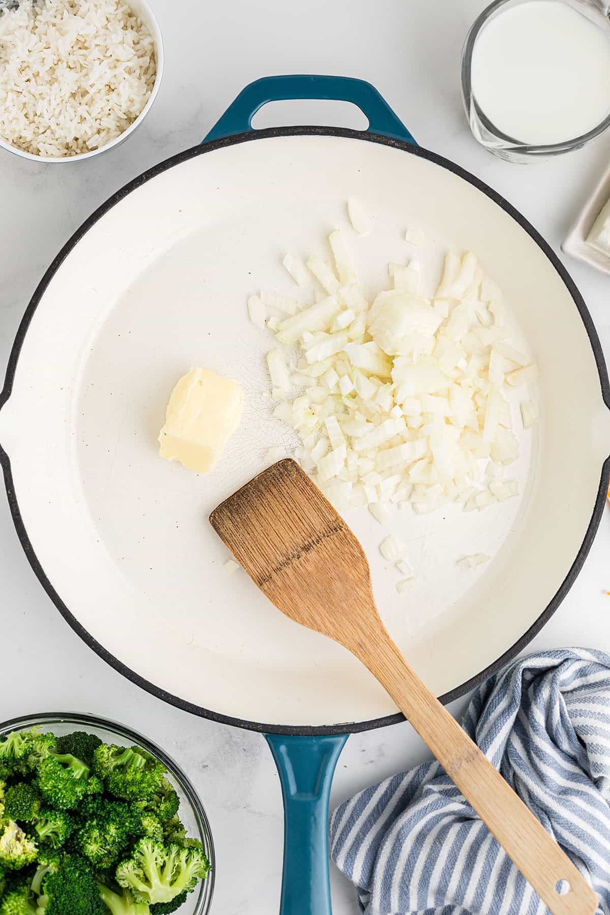 Chopped onions and butter in a skillet with a wooden spatula, broccoli and rice in bowls nearby.