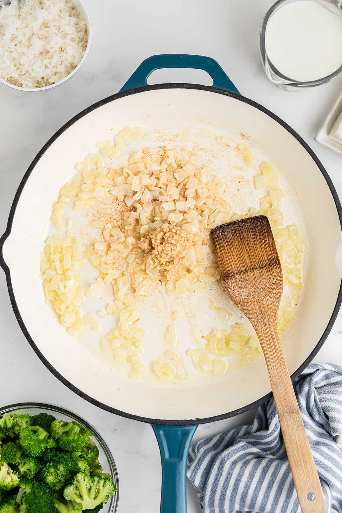 Chopped onions and garlic sautéing in a white skillet with a wooden spatula, broccoli and rice nearby.