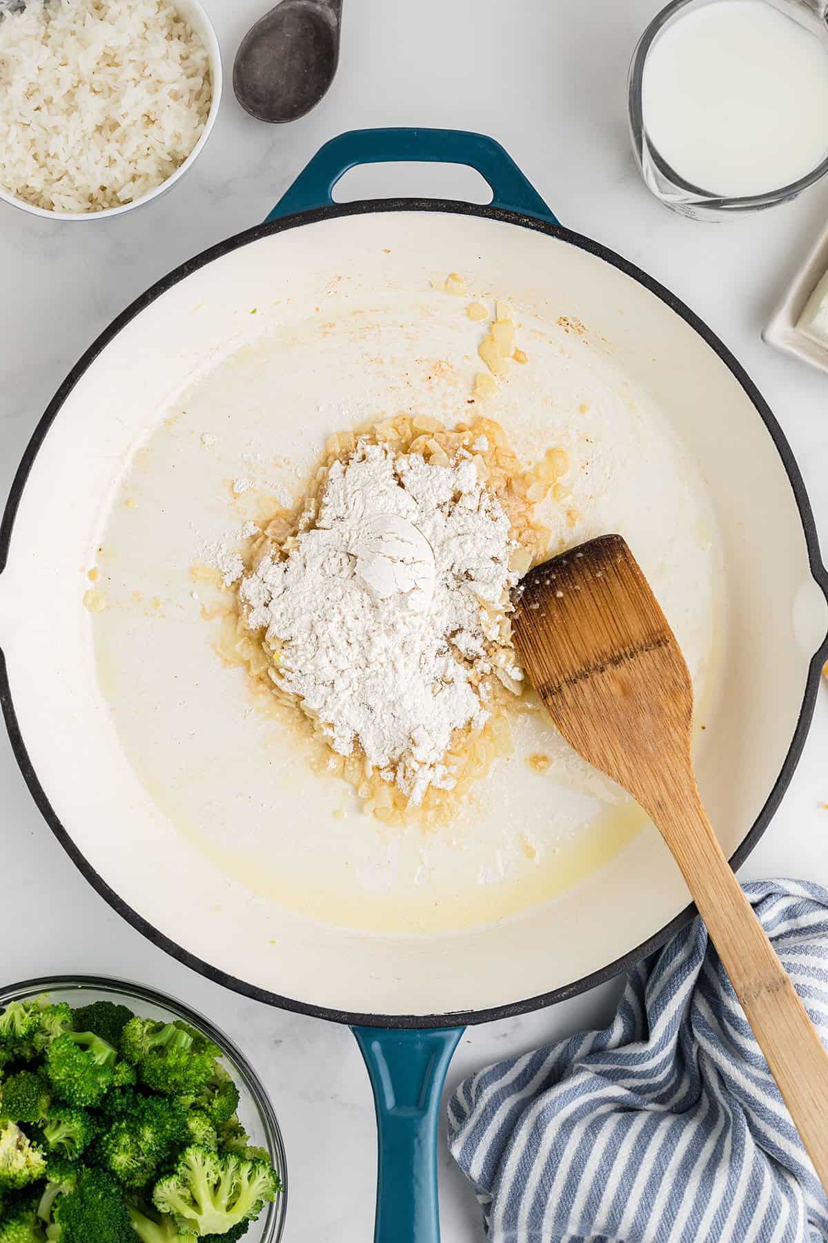 Flour and onions cooking in a white pan with a wooden spatula, surrounded by broccoli, milk, and rice.