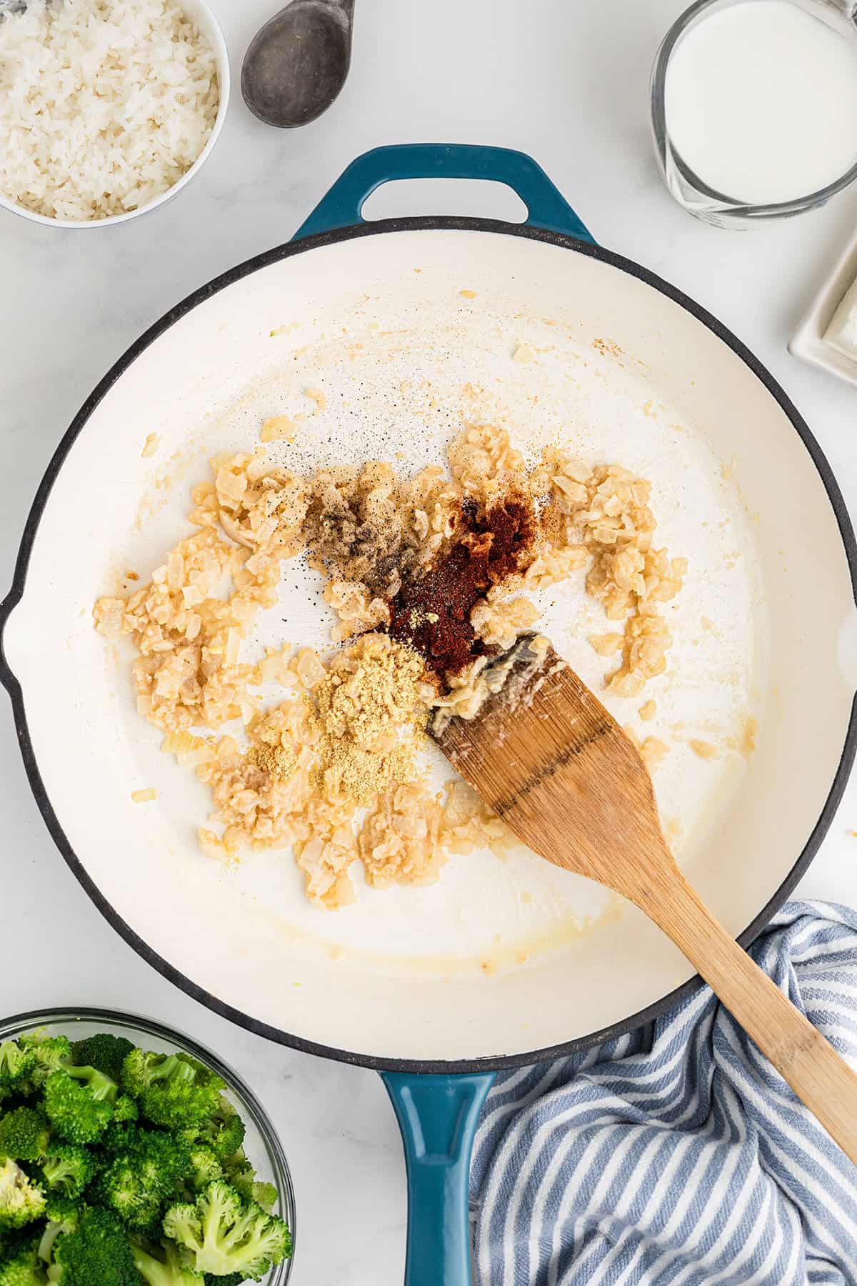 A skillet with sautéed onions, spices, and a wooden spatula; bowls of rice, milk, and broccoli nearby.