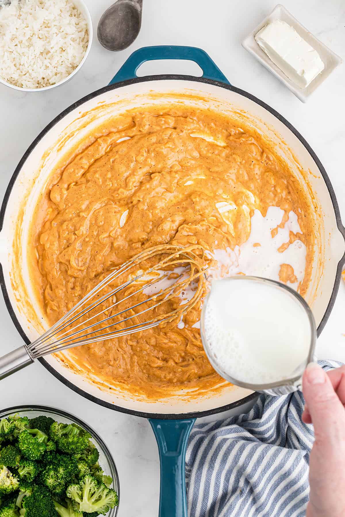 A hand pours milk into a pan of sauce being whisked, with rice, broccoli, and butter nearby.