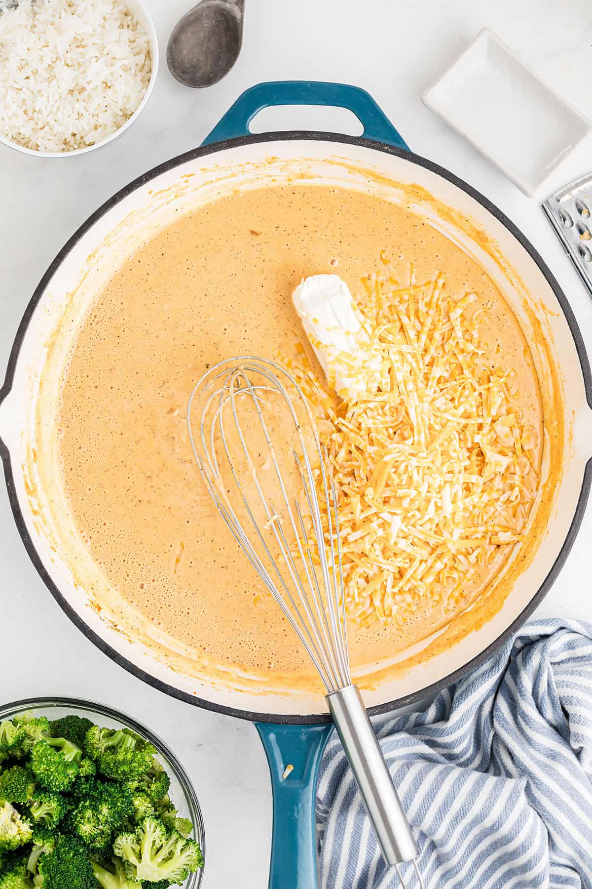 A whisk stirs shredded cheese and cream into a creamy sauce in a skillet, surrounded by rice and broccoli.