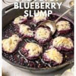 A skillet filled with blueberry slump topped with baked dumplings, surrounded by dishes and blueberries.