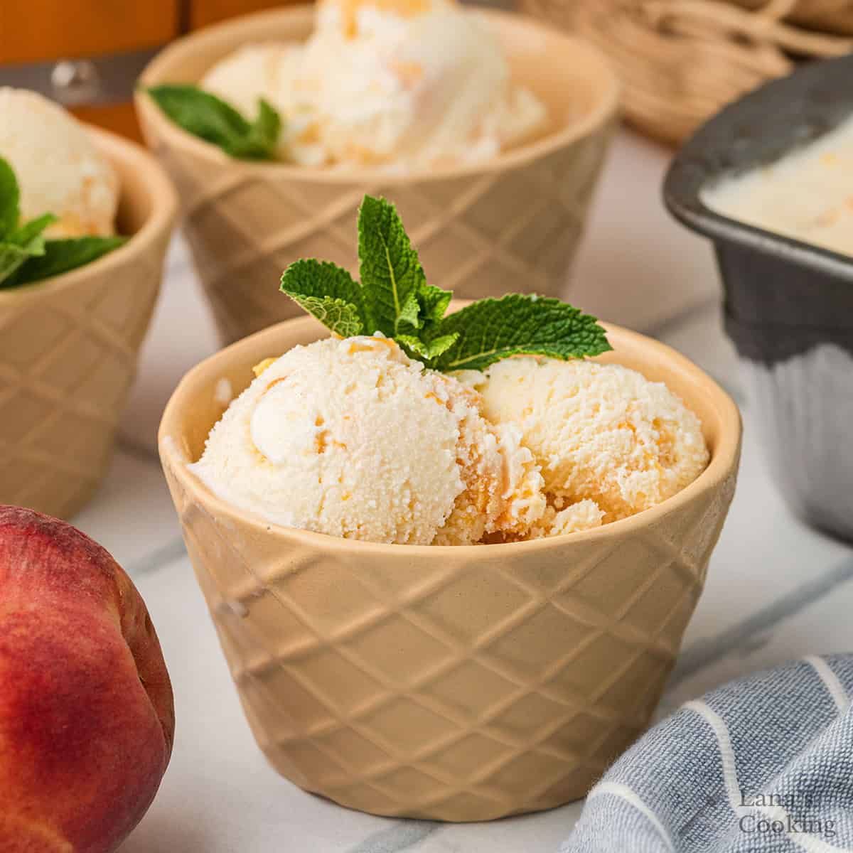 A beige bowl with a waffle pattern holds two scoops of vanilla ice cream, garnished with a sprig of mint. A fresh peach and another bowl of ice cream are nearby, with a striped cloth partially visible.