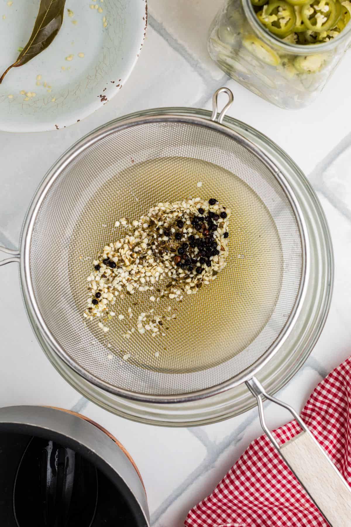 A strainer over a jar with the garlic and peppercorns strained from the brine.