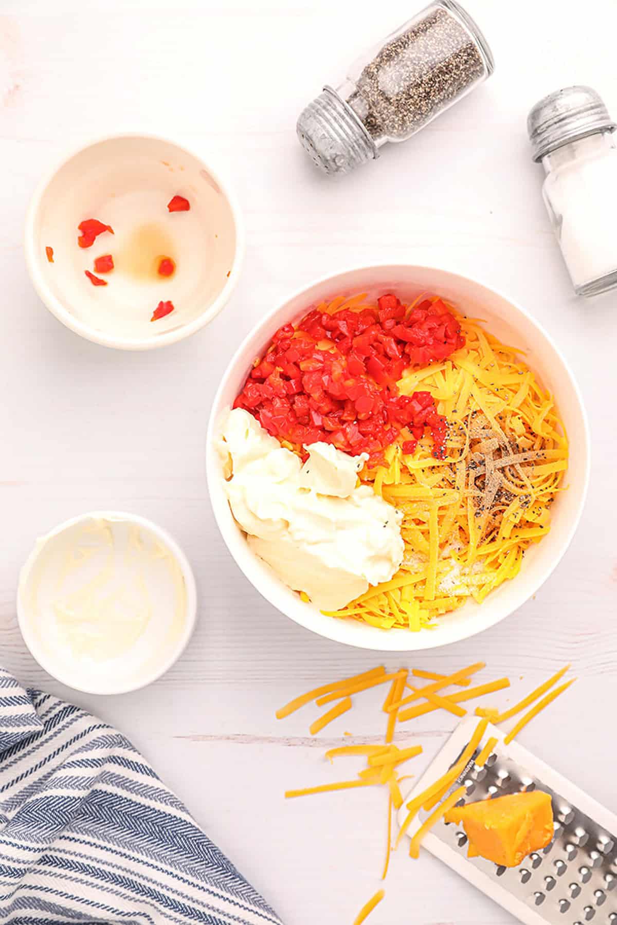 Bowl with shredded cheese, diced peppers, mayonnaise, and seasoning on a white table with salt and pepper shakers.