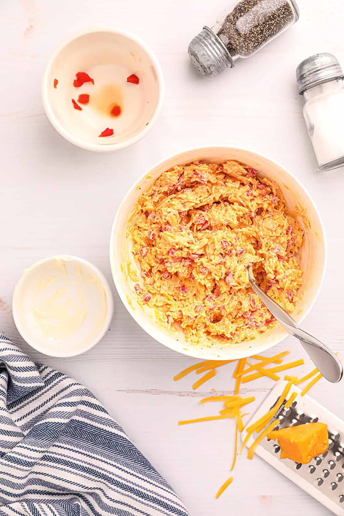Bowl of pimiento cheese spread with a spoon, grated cheese, seasoning, and empty bowls on a white surface.