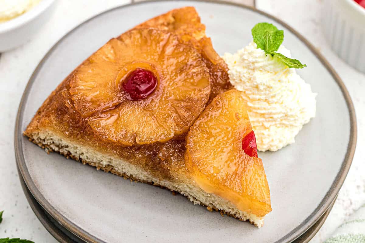 A slice of pineapple upside down cake on a plate with whipped cream on the side.