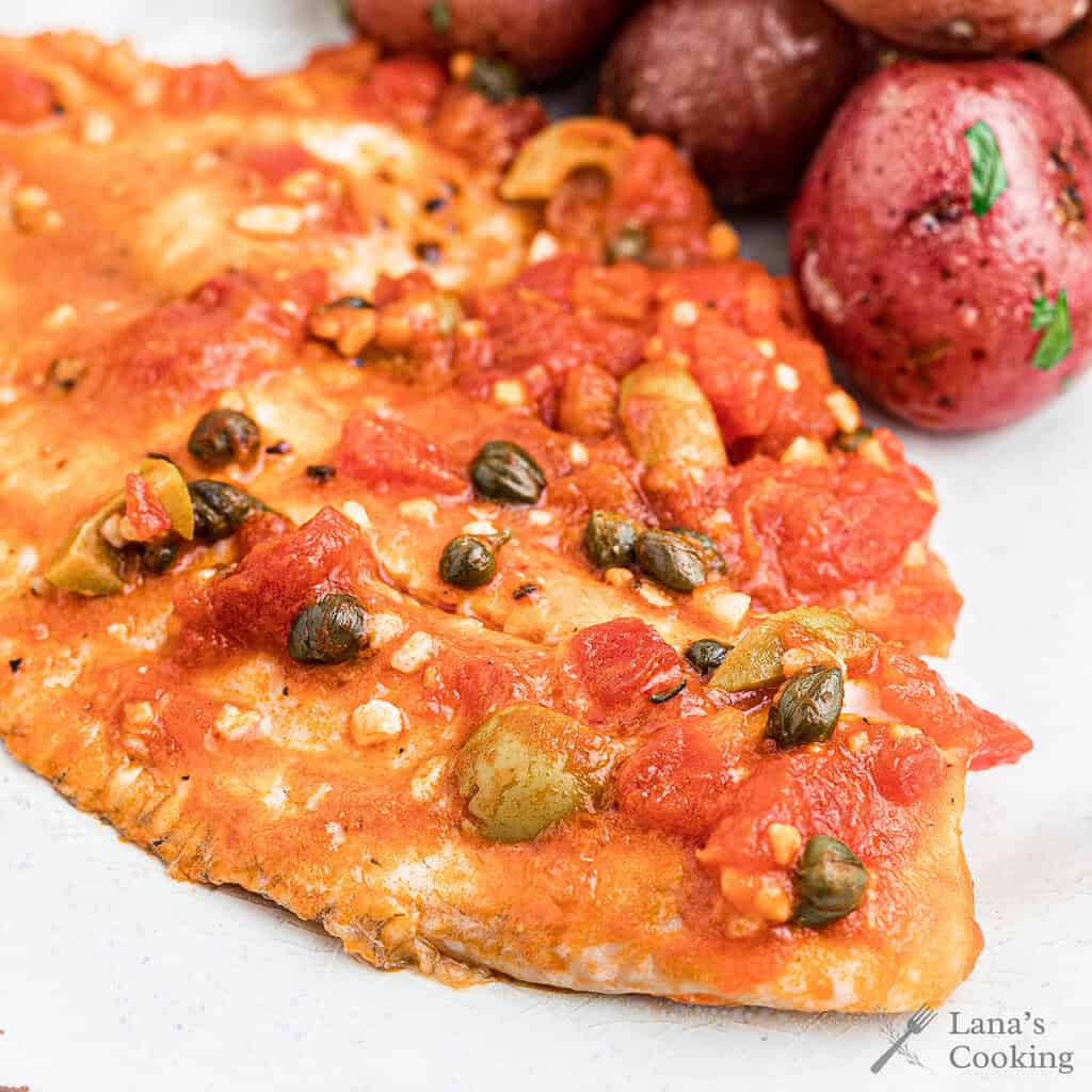 Fish fillet with tomato, olives, and capers, served alongside red potatoes on a white plate.