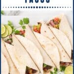 Six carne asada tacos in flour tortillas with lime slices and cilantro on a white tray.