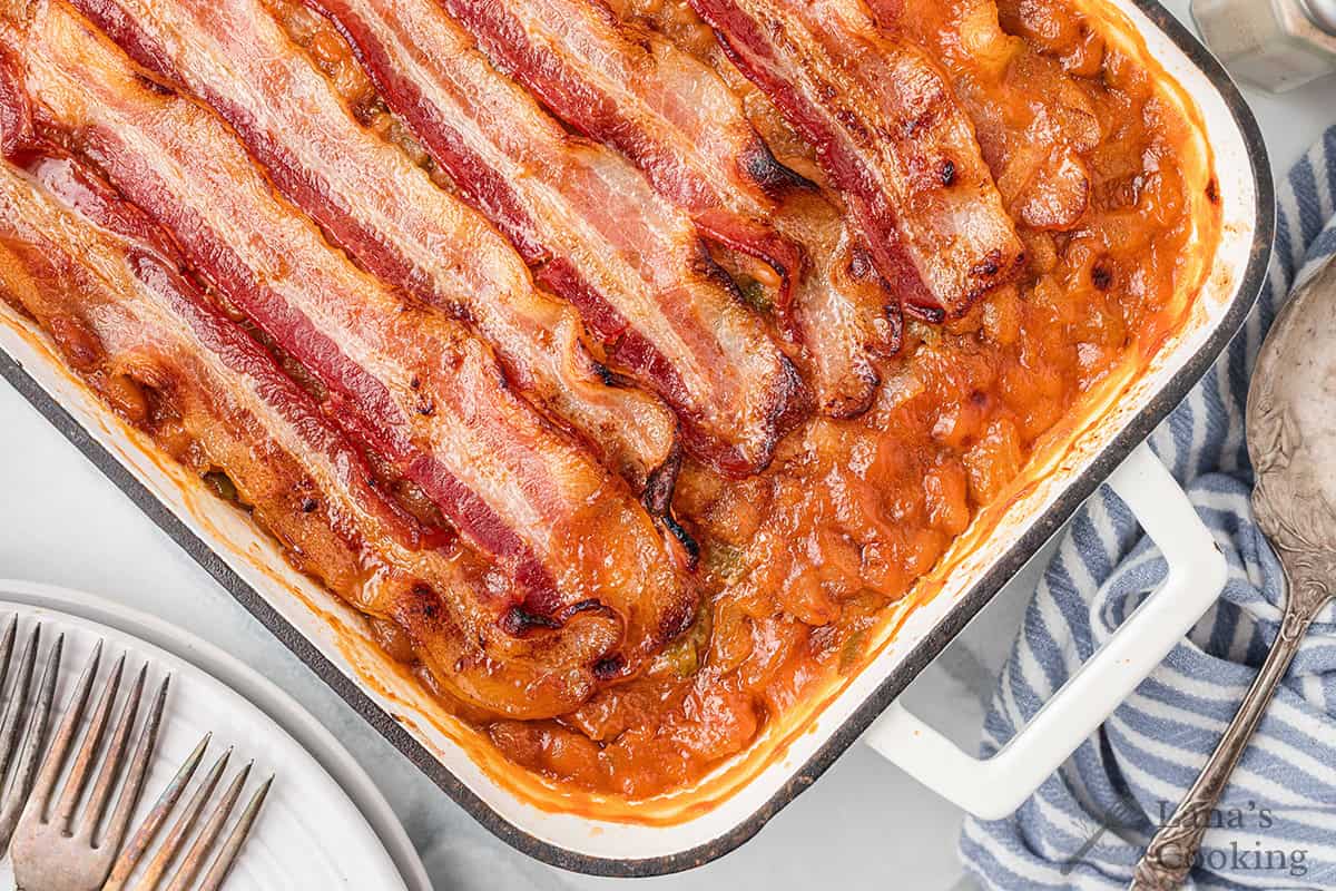 A baking dish filled with baked beans topped with strips of crispy bacon, next to plates and utensils.