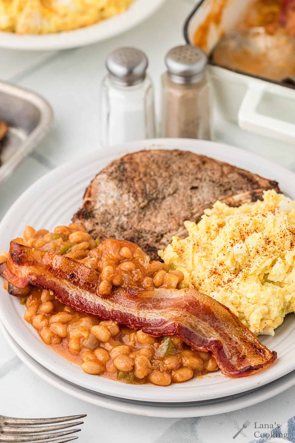 A plate with potato salad, baked beans topped with bacon, and a steak.