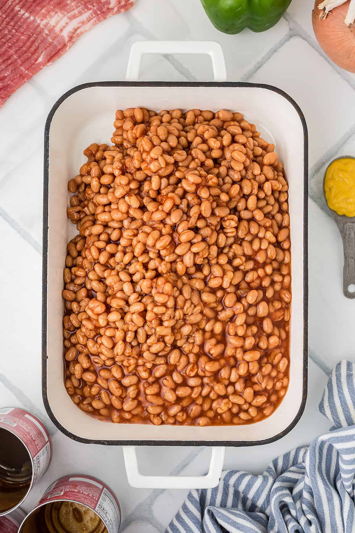 A white baking dish filled with pork and beans, surrounded by cans, bacon, a bell pepper, and a spoon of mustard.