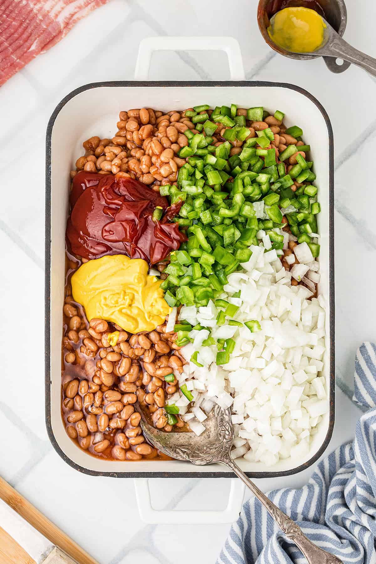 Rectangular dish with beans, ketchup, mustard, chopped green bell pepper, and chopped onions, unmixed.