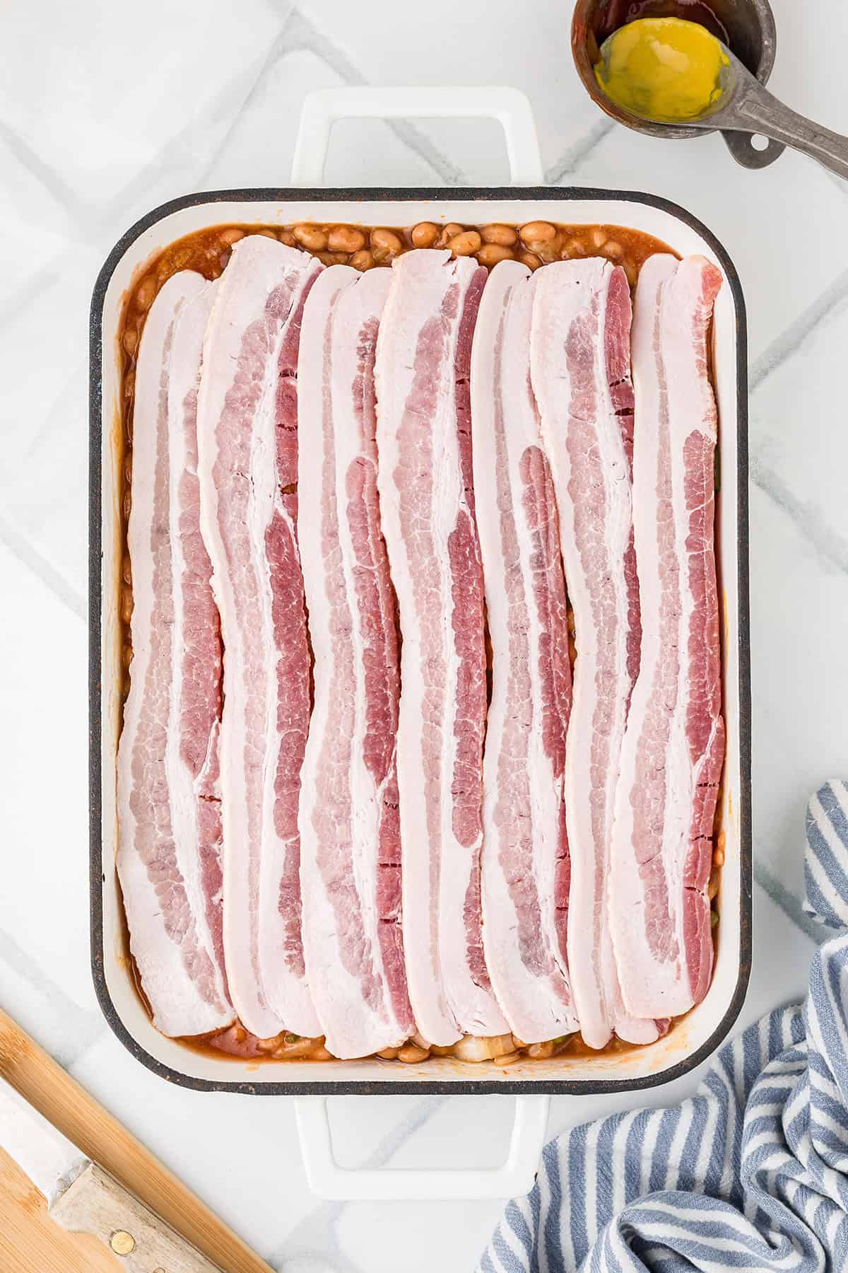 Uncooked bacon strips layered over beans in a rectangular white casserole dish on a marble surface.