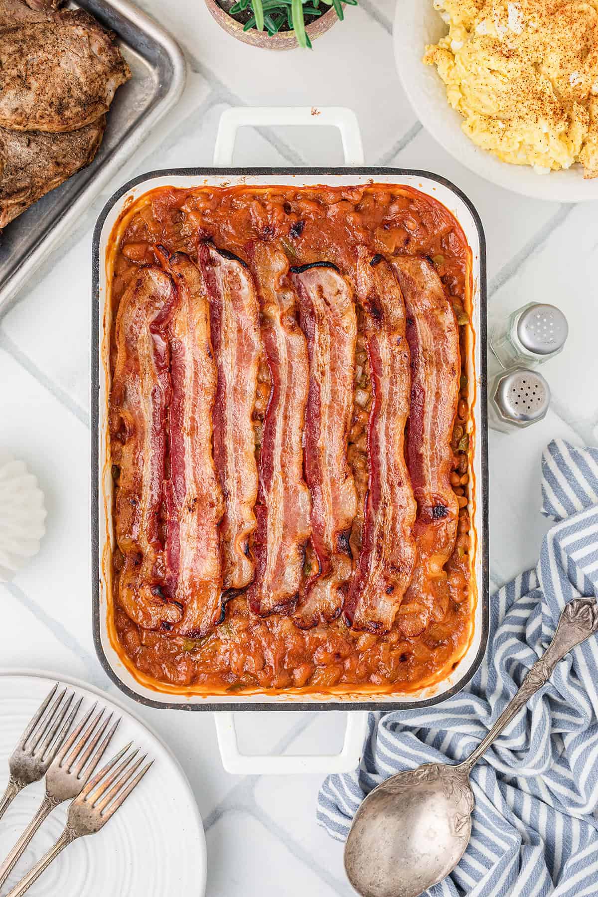 A casserole dish of baked beans topped with bacon strips, surrounded by serving utensils and side dishes.