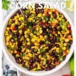 A bowl of black bean and corn salad with a spoon, surrounded by fresh lime and tomato.