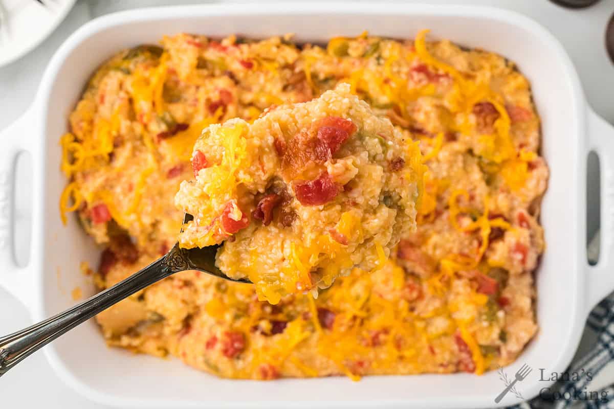 A spoonful of cheesy tomato grits with tomatoes is held above a white baking dish filled with the casserole.