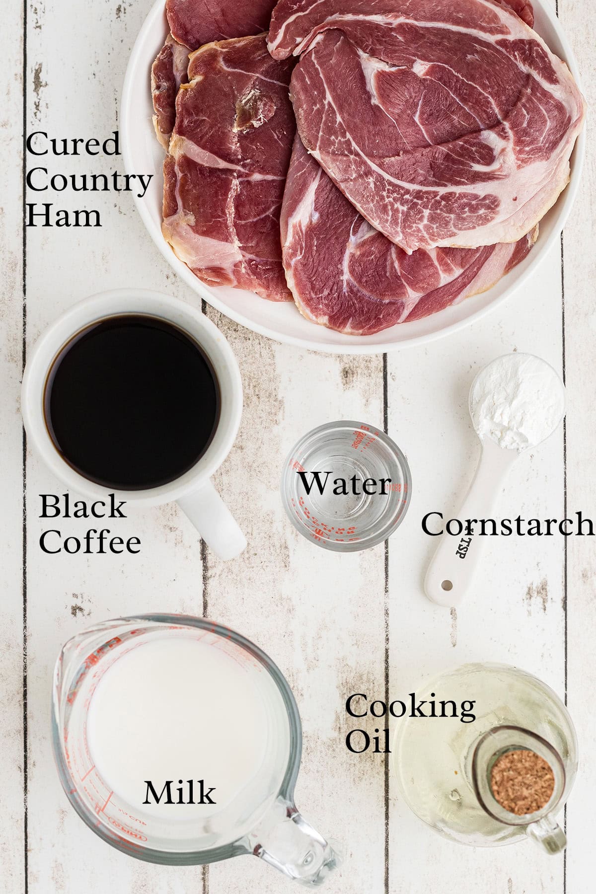 A bowl of cured country ham slices, a cup of black coffee, a small glass of water, a measuring spoon of cornstarch, a measuring cup of milk, and a small pitcher of cooking oil on a white wooden surface.