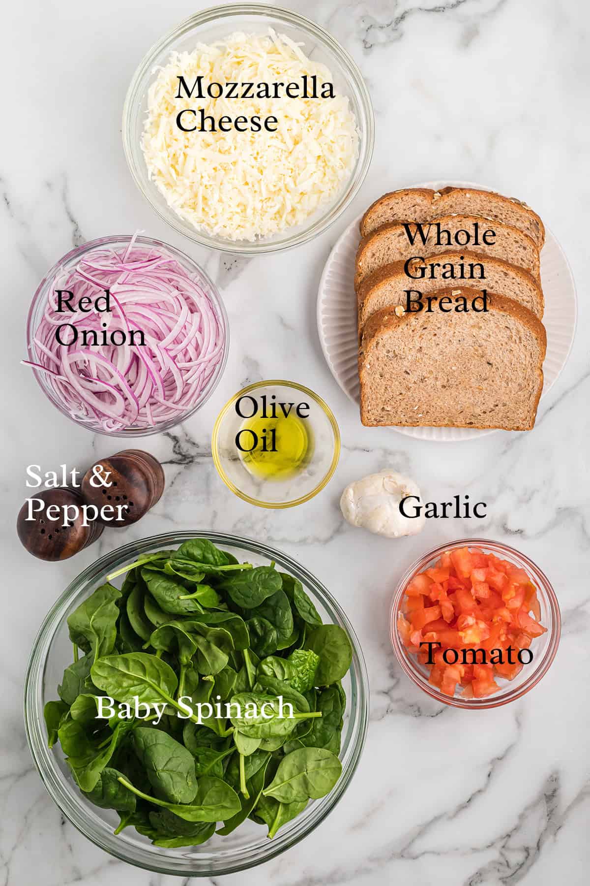 Ingredients on a marble counter: mozzarella, whole grain bread, red onion, olive oil, garlic, tomato, spinach, salt, pepper.