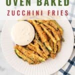 A white plate with breaded zucchini fries arranged next to a small bowl of white dipping sauce, placed on a blue and white cloth napkin. Text on the image reads, Oven Baked Zucchini Fries, and lanascooking.com.