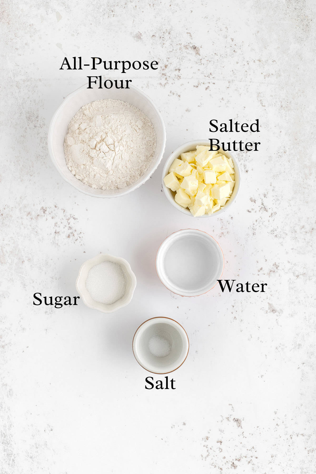 A flat lay of five bowls containing labeled ingredients: all-purpose flour, salted butter, water, sugar, and salt, placed on a white textured surface.