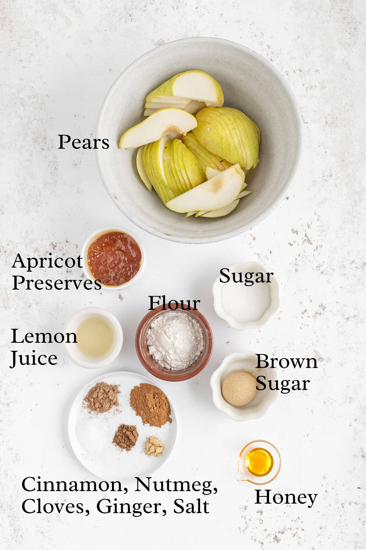 A bowl of sliced pears surrounded by small bowls containing apricot preserves, sugar, brown sugar, lemon juice, flour, honey, and spices labeled cinnamon, nutmeg, cloves, ginger, and salt on a white surface.