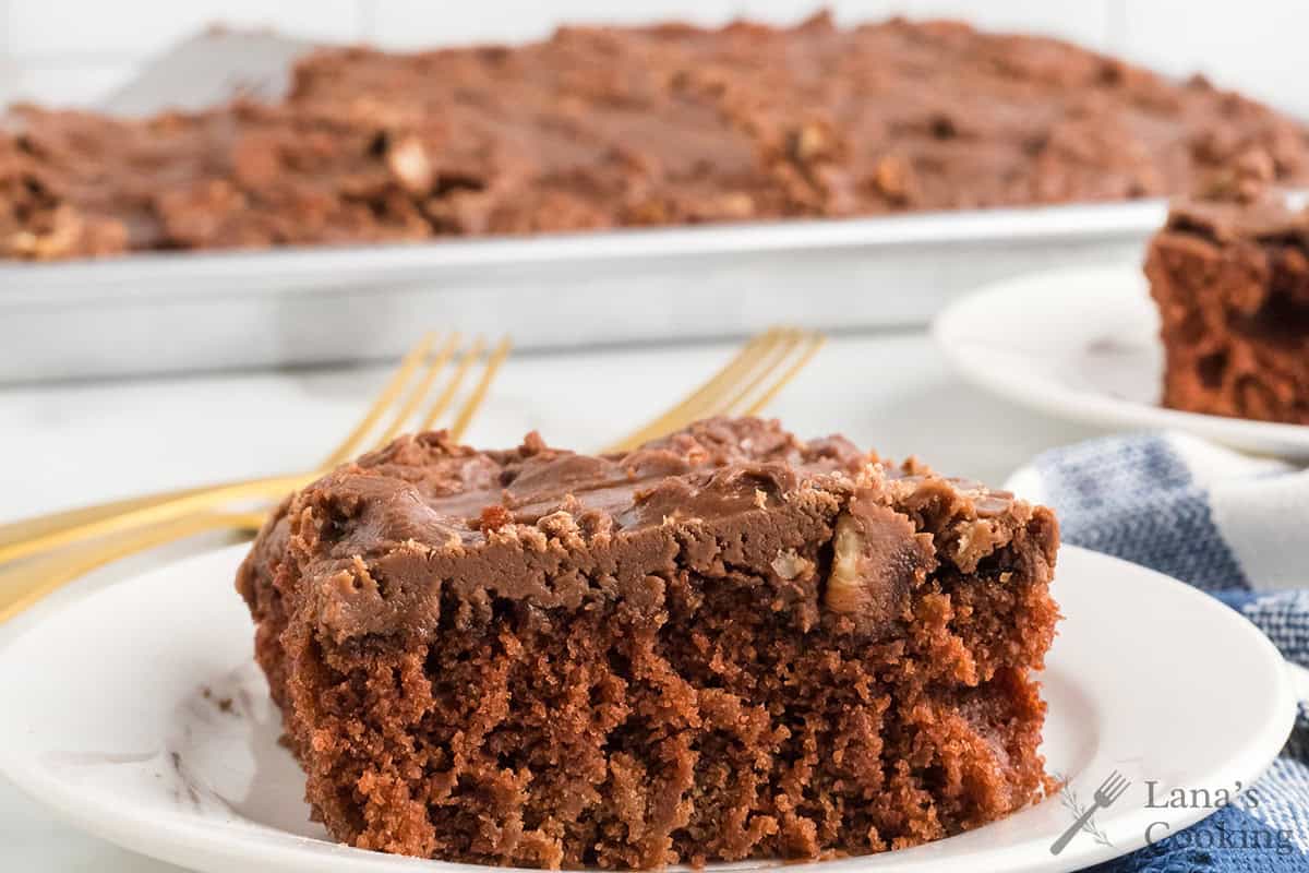 A slice of chocolate sheet cake with chocolate frosting sits on a white plate beside gold forks.