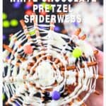 A hand holds a white chocolate pretzel spiderweb decorated with candy spiders and colorful candies.