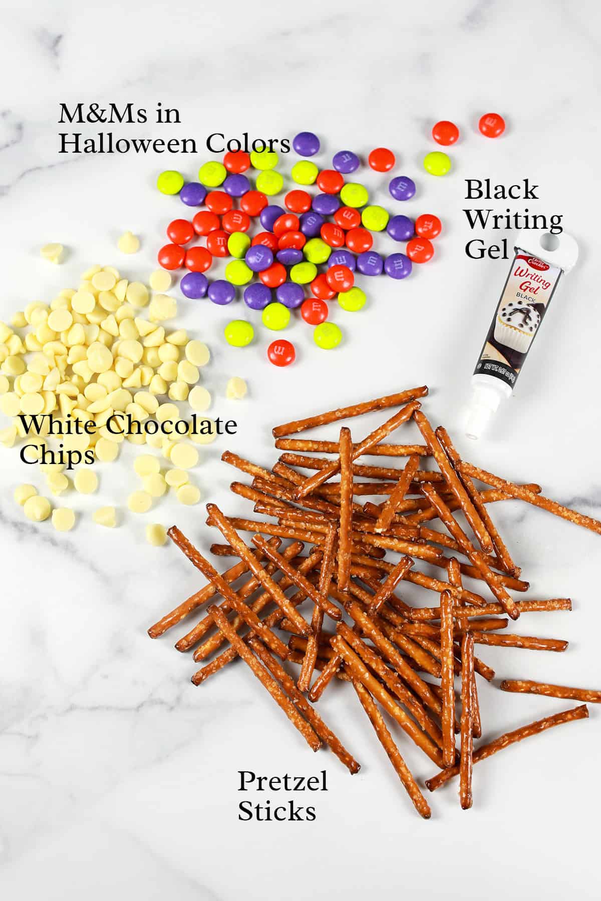 M&Ms, white chocolate chips, black writing gel, and pretzel sticks arranged on a white marble surface.