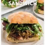 Steak sandwich with grilled onions and arugula on a plate, topped with a ciabatta roll.