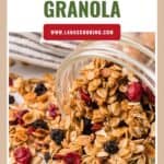 A jar of homemade granola with oats, dried berries, and nuts spilling onto a white surface.