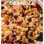 Granola with oats, nuts, and dried berries spills from a glass jar onto a white surface.