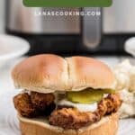 Crispy fried chicken sandwich with pickles and sauce on a bun, served on a white plate.