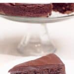 A slice of flourless chocolate cake with ganache on a plate; whole cake in the background.